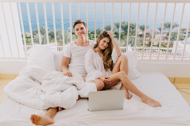 Free photo handsome young man in white shirt smiling while his pretty girlfriend dreamy touching her long hair. beautiful couple spending time together with laptop posing