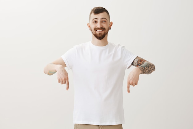 Free photo handsome young man with tattoos pointing fingers down at announcement