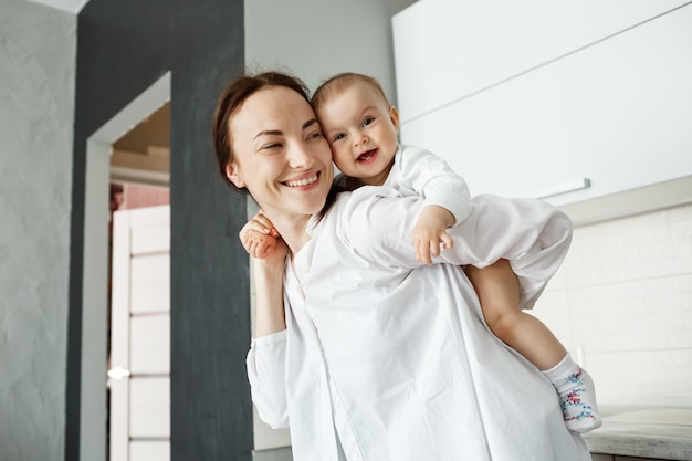 Handsome young mother playing with little baby boy holding him on back with hands. Child brightfully smiling and feeling happy.