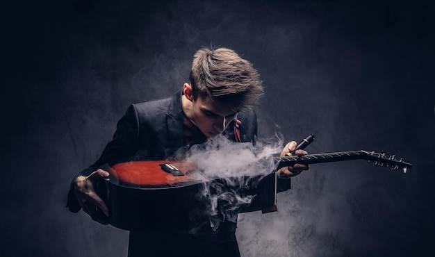 Free photo handsome young musician with stylish hair in elegant clothes exhales smoke on his acoustic guitar. isolated on a dark background.