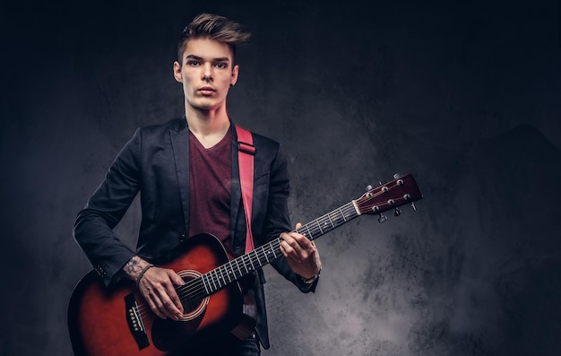 Handsome young musician with stylish hair in elegant clothes with a guitar in his hands playing and posing on a dark background.