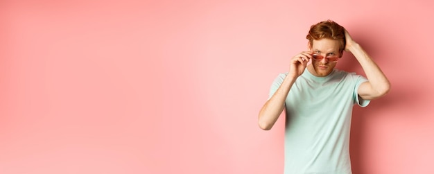 Free Photo handsome young redhead man in sunglasses brushing hair with hand and looking smug and confident at c