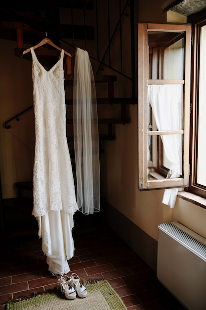 Free photo hanging classy wedding dress and wedding veil in the room near the opened window