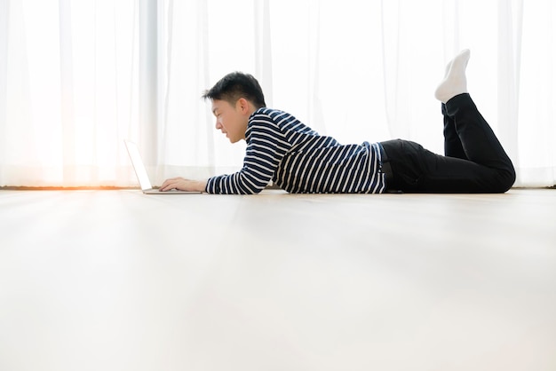 Free photo happiness asian smart male smile with confident and joyful working with laptop casual tshirt relax and lay down on wooden floor with home window interior background