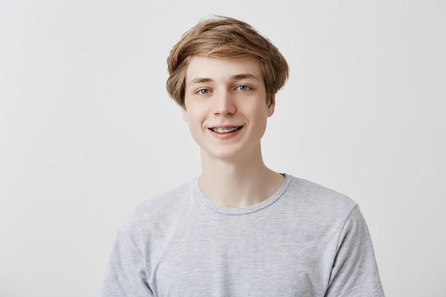 Happiness and positive human expressions. Studio shot of friendly young male student with fair hair and blue eyes laughing at good joke, smiling with braces, looking  with carefree smile