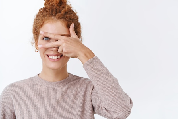 Free photo happiness wellbeing and lifestyle concept gorgeous feminine lovely redhead woman with curly hair combed in bun wear stylish blouse showing peace sign as cover face with hand smiling