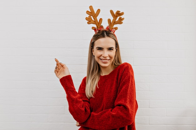 Happy adorable lady in red pullover in christmas headgear smiling and looking at camera over isolated background