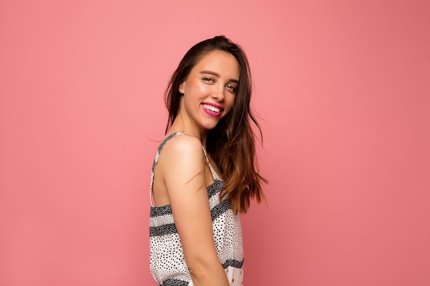 Happy adorable lady with long wavy hair in summer dress posing over pink ackground