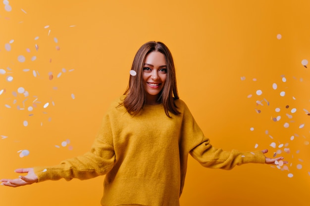 Free photo happy adorable woman in yellow sweater laughing on bright wall. indoor portrait of appealing girl posing with confetti.