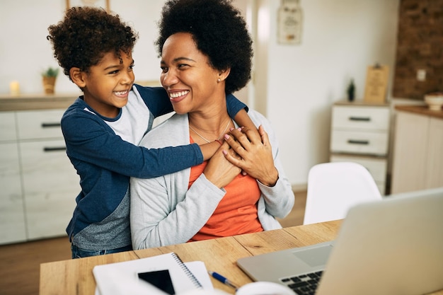 Happy African American boy embracing his mother who is working on laptop at home