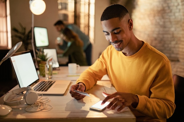 Free photo happy african american entrepreneur using mobile phone and credit card from online payments while working at night in the office