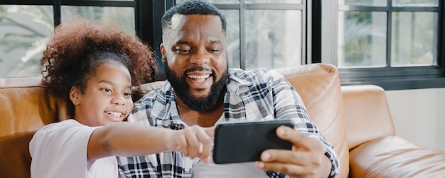 Happy African American family dad and daughter having fun and using mobile phone video call on sofa at house.