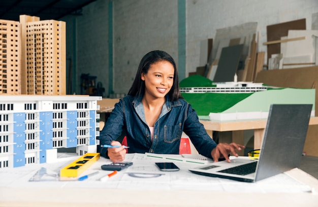 Happy African-American lady with laptop and model of building at table