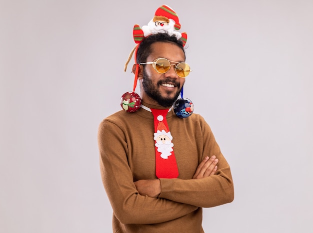 Free photo happy african american man in brown sweater and santa rim on head with funny red tie holding christmas balls looking at camera with smile on face standing over white background