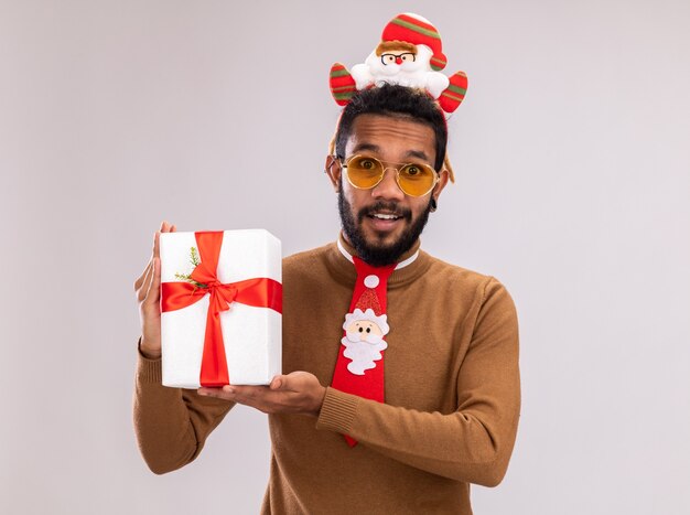 Happy african american man in brown sweater and santa rim on head with funny red tie holding a present looking at camera with smile on face standing over white background