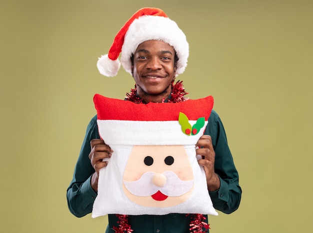 Happy african american man in santa hat with garland holding christmas pillow looking at camera with smile on face standing over green background