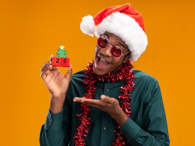 Happy african american man in santa hat with garland wearing sunglasses holding toy cubes with date twenty five presenting with arm standing over orange background