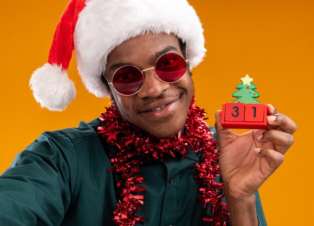 Happy african american man in santa hat with garland wearing sunglasses holding toy cubes with new year date  smiling standing over orange wall