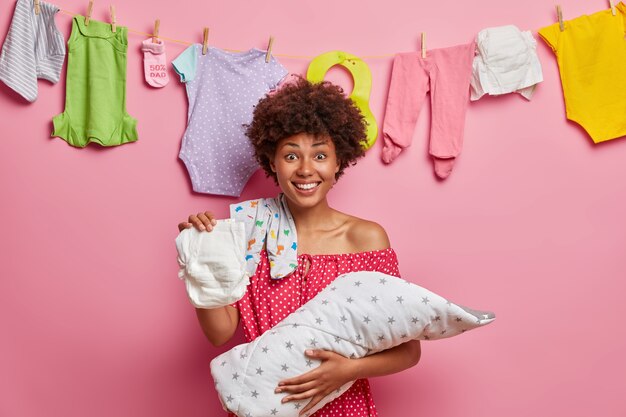 Happy African American mother cares about infant, changes nappy on baby daughter, plays with child, stands indoor at home, poses 