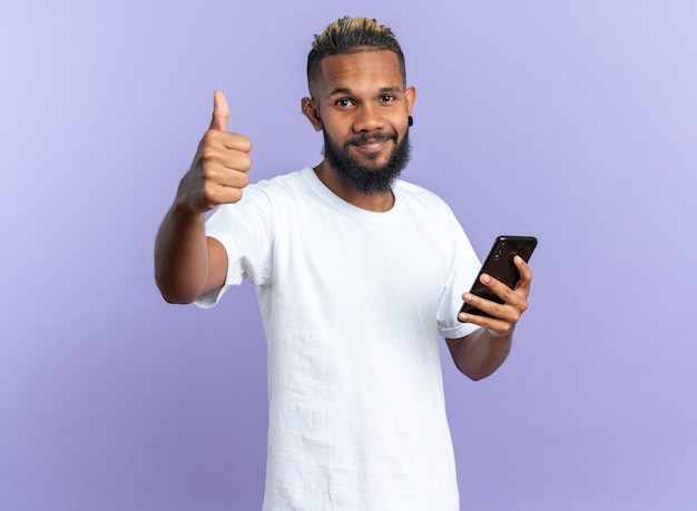 Happy african american young man in white t-shirt holding smartphone looking at camera showing thumbs up smiling cheerfully