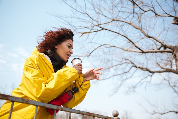 Free photo happy african curly young woman wearing yellow coat drinking coffee.