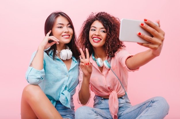 Happy african girl with pretty face posing with peace sign near charming female friend. Aforable mulatto woman in jeans and pink shirt making selfie with stylish hispanic lady.