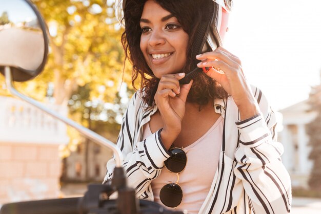 happy african woman sitting on modern motorbike and fastens her moto halmet outdoors