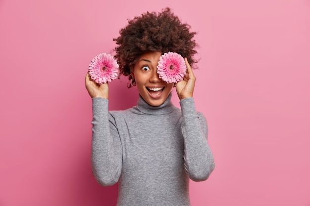 Free photo happy afro american woman covers eyes with rosy gerbera flowers, has fun and giggles positively, going to decorate room for party