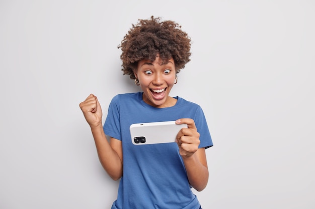 Happy Afro American woman dressed in casual blue t shirt holds smartphone horizontally clenches fist wins online watches video isolated over white wall