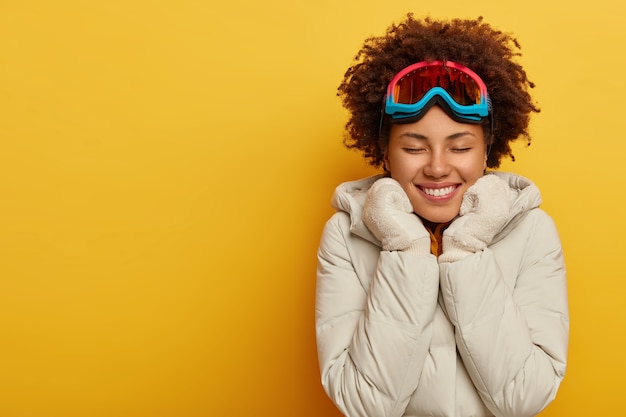 Happy Afro woman wears ski mask, warm knitted mittens and coat, enjoys outdoor activities. Glad female in snowboard wear