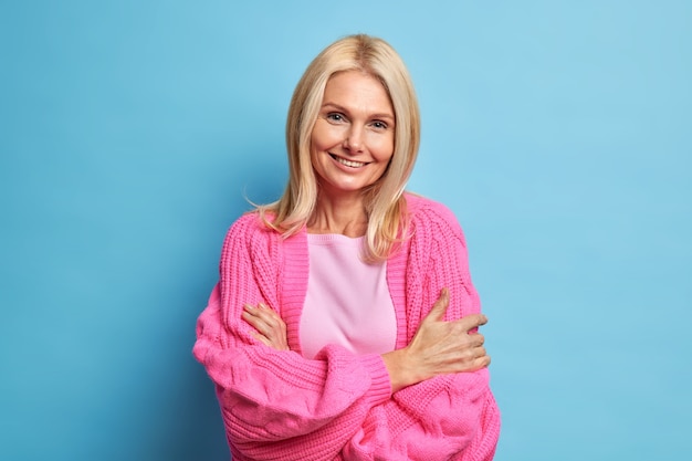 happy aged grandmother looks with gentle smiles keeps arms folded stands in relaxed pose dressed in casual knitted sweater.