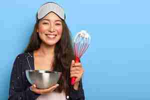 Free photo happy asian brunette woman makes delicious cake, prepares cake, whisks egg white in bowl with beater, dressed in nightwear, sleep mask