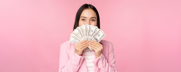 Happy asian lady in suit holding money dollars with pleased face expression standing over pink background