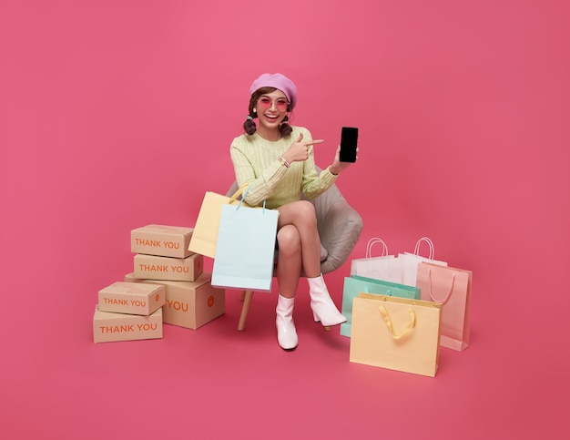 Happy Asian teen woman sitting on sofa holding shopping bags and smartphone isolated on pink background Shopper or shopaholic concept