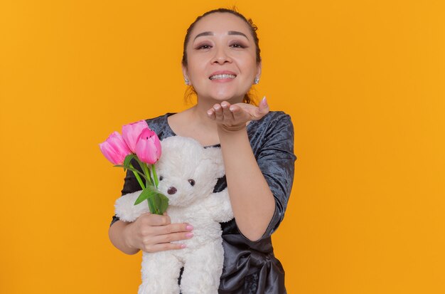 happy asian woman holding bouquet of pink tulips and teddy bear blowing a kiss