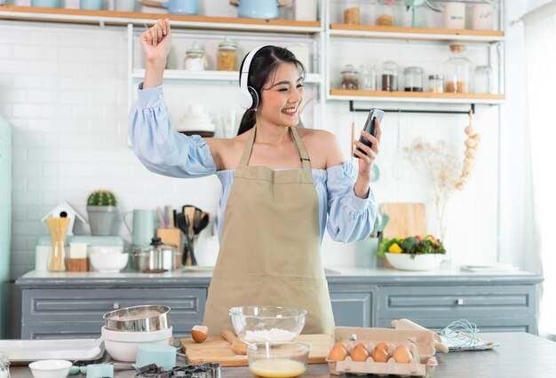 Happy asian woman raise smartphone fist good mood wear headphones listen to music in kitchen at home