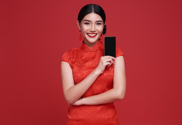 Happy Asian woman wearing traditional cheongsam qipao dress showing mobile phone isolated on red background.