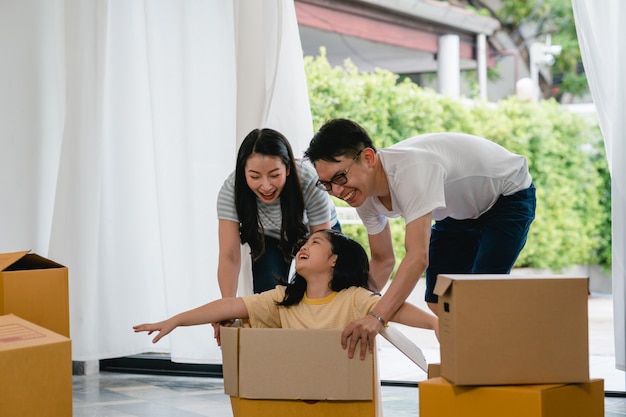 Happy Asian young family having fun laughing moving into new home. Japanese parents mother and father smiling helping excited little girl riding sitting in cardboard box. New property and relocation.