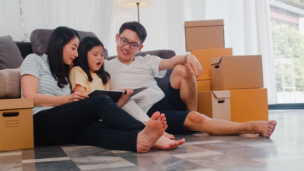 Happy Asian young family homeowners bought new house. Chinese Mom, Dad, and daughter embracing looking forward to future in new home after moving in relocation sitting on floor with boxes together.