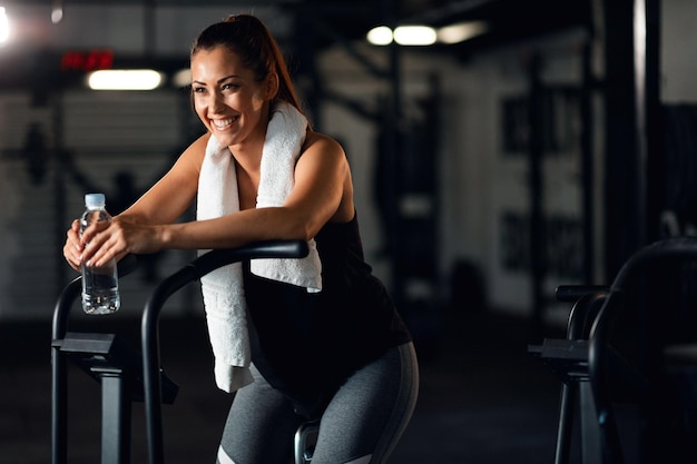 Free photo happy athletic woman with water bottle taking a break and resting on exercise bike after cycling in a gym copy space