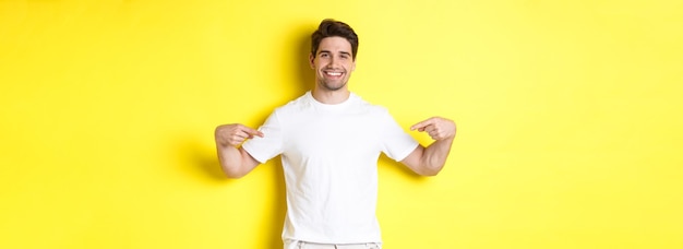 Free photo happy attractive guy pointing fingers at your logo showing promo on his tshirt standing over yellow