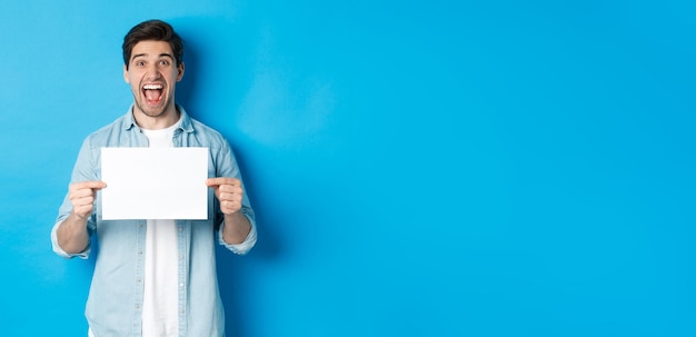 Free photo happy attractive man showing piece of paper for your logo sign standing amazed against blue backgrou