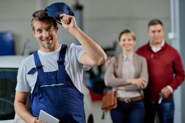 Free Photo happy auto mechanic taking his hat of while standing in a workshop and looking at camera his customers are in the background