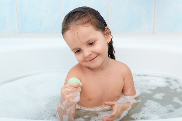 Happy baby taking bath alone, playing with foam bubbles and her new toy.