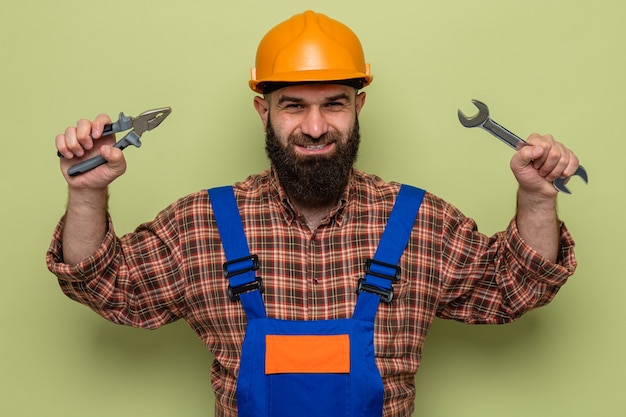 Free Photo happy bearded builder man in construction uniform and safety helmet holding wrench and pliers looking smiling cheerfully