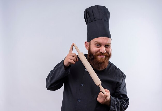 A happy bearded chef man in black uniform showing rolling pin while looking at camera on a white wall