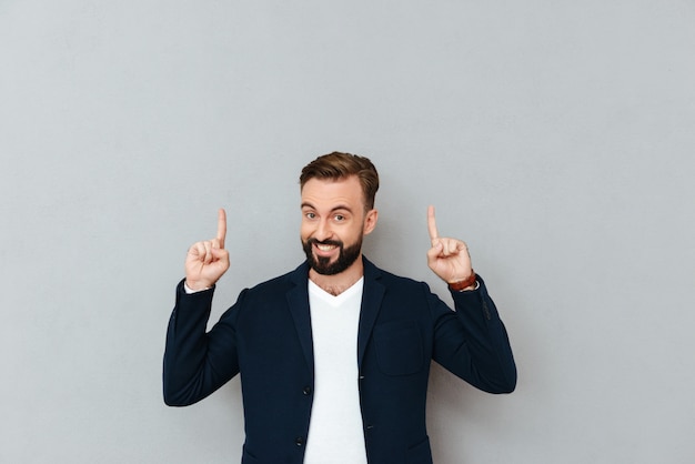 Happy bearded man in busines clothes pointing up and looking at the camera over gray 