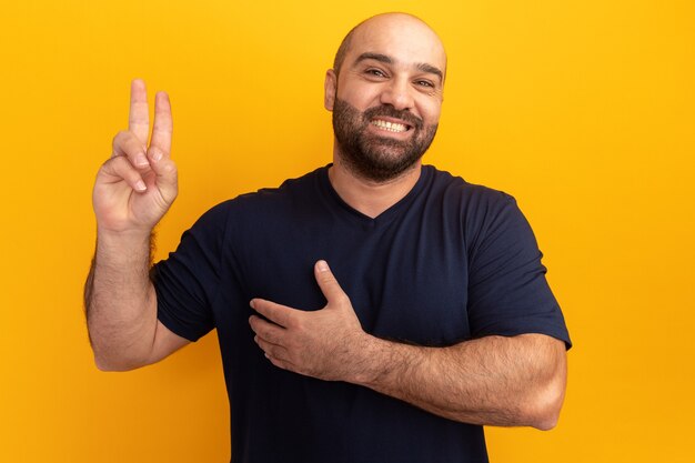 Happy bearded man in navy t-shirt holding hand on chest showing fingers making a promise standing over orange wall