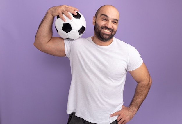 Happy bearded man in white t-shirt holding soccer ball on his shoulder smiling standing over purple wall