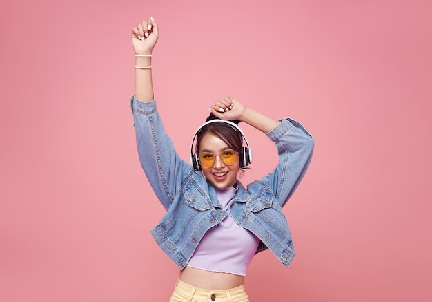 Free Photo happy beautiful asian teen woman in yellow glasses listening music in headphones and dance on pink wall.